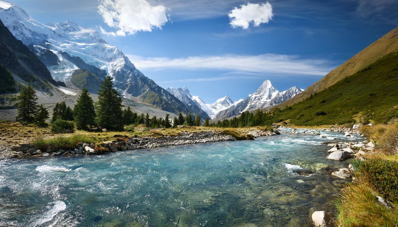 Stunning mountain view with clear river, snow-capped peaks, and lush greenery in summer.