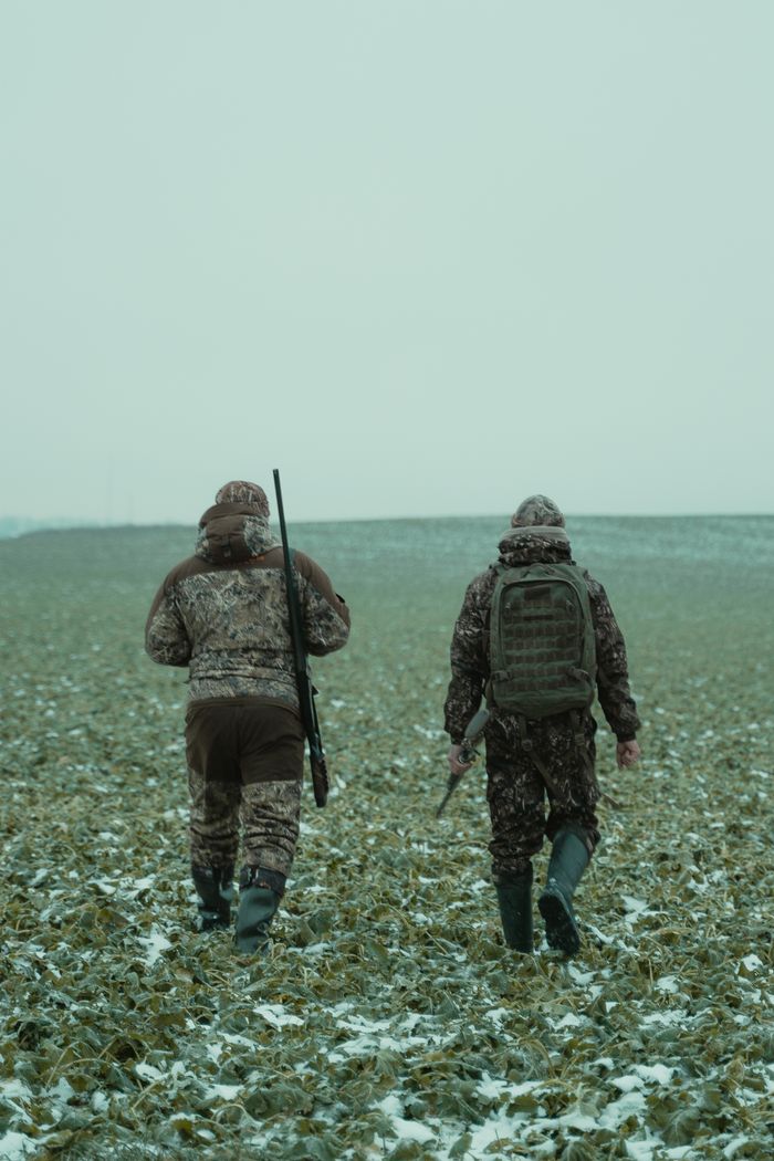 Two hunters in camouflage gear walk across a snowy field, back view.